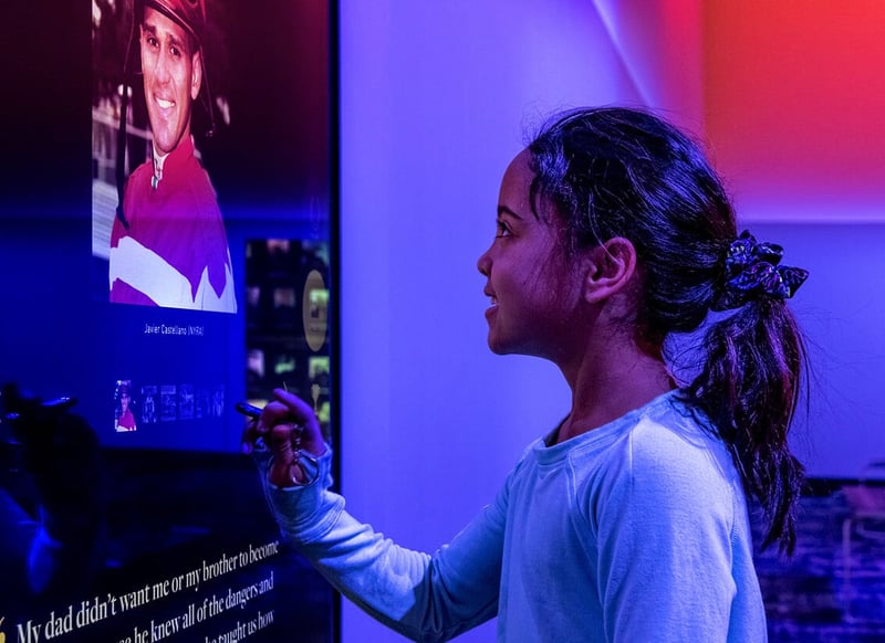 A young girl interacting with a touchscreen at the National Racing Museum and Hall of Fame. The screen displays an image of jockey Javier Castellano, and below it is a quote that begins with “My dad didn’t want me or my brother to become...” — suggesting a personal story of perseverance or inspiration.