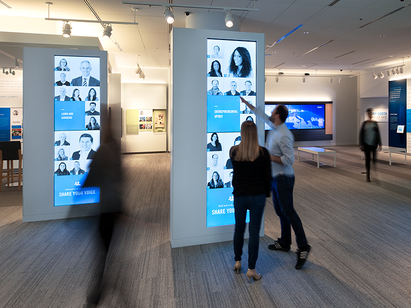 An interactive museum exhibit showing tall vertical digital screens embedded in freestanding white pillars. The screens display photos and brief text about various people, with themes like 
