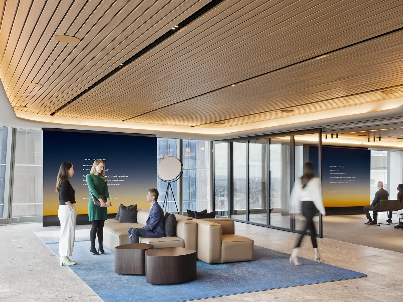 A modern, high-rise corporate office lobby with floor-to-ceiling windows offering a city view. The space features wooden slatted ceilings with ambient lighting, contemporary leather seating, and minimalist round coffee tables on a blue rug. Three professionals are engaged in conversation near the seating area, while two others are seated in the background. A woman is walking through the space, which has a clean and airy atmosphere. Digital display panels on the walls show gradient backgrounds with white text, enhancing the sleek and professional setting.