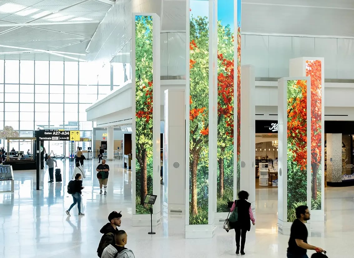 A modern airport terminal interior with travelers walking through. Tall digital display columns show vivid images of trees with green and red foliage, creating a vibrant forest-like illusion within the space. Gates A27–A34 are visible in the background.