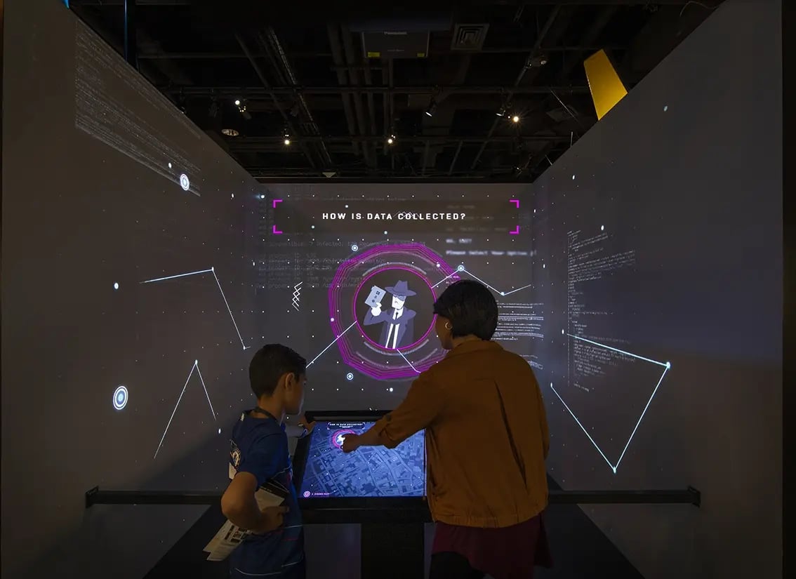 A woman and a boy interact with a digital touchscreen in a dark exhibition space. The screen and walls display a futuristic interface with neon graphics and the question 