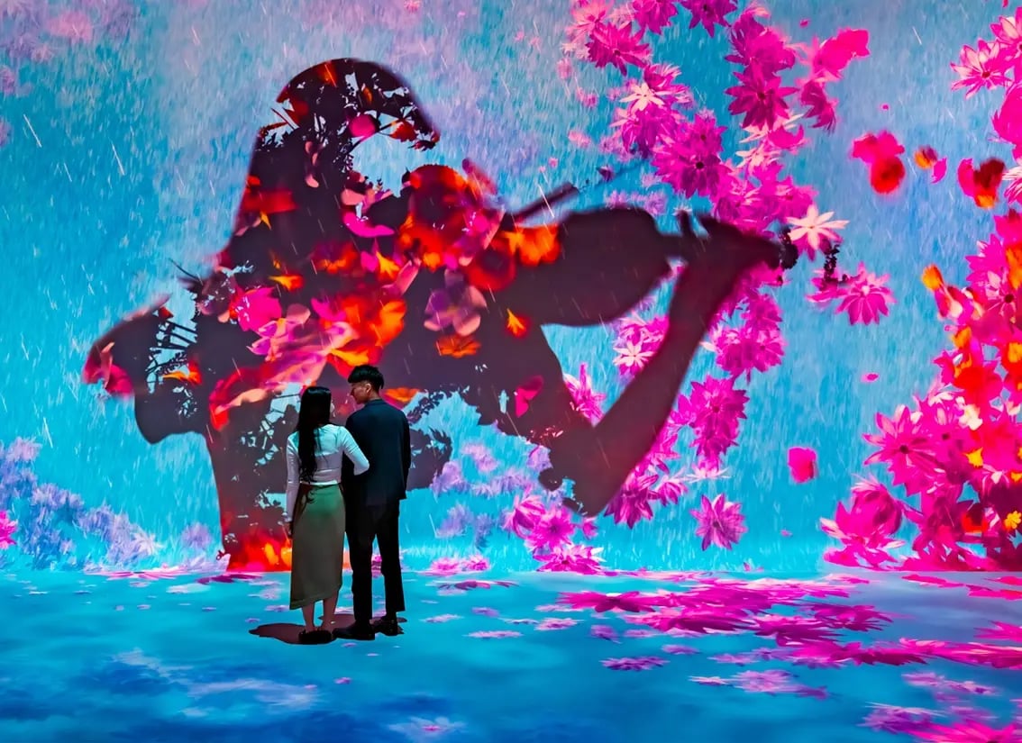 A couple stands closely together, gazing at a vibrant digital art installation. The immersive environment surrounds them with vivid colors—shades of blue, pink and orange—depicting falling petals and a large silhouette of a violinist. The scene evokes a dreamy, romantic atmosphere with floral projections covering the walls and floor.