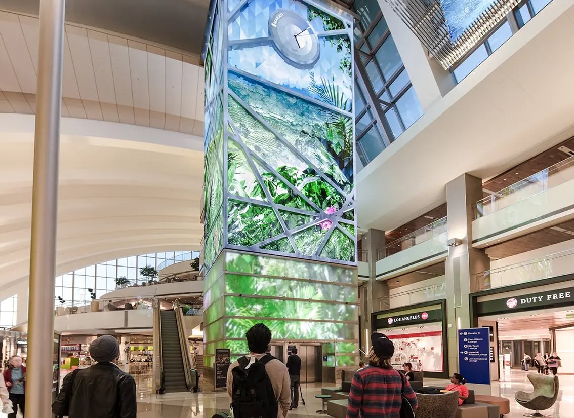 Travelers look up at a towering digital installation in a spacious, modern airport terminal. The column features vivid, nature-themed visuals such as lush greenery and landscapes, framed by geometric lines and topped with a circular clock. Surrounding the installation are shops, including a duty-free store, escalators and comfortable seating areas. Natural light floods the area from large windows above.